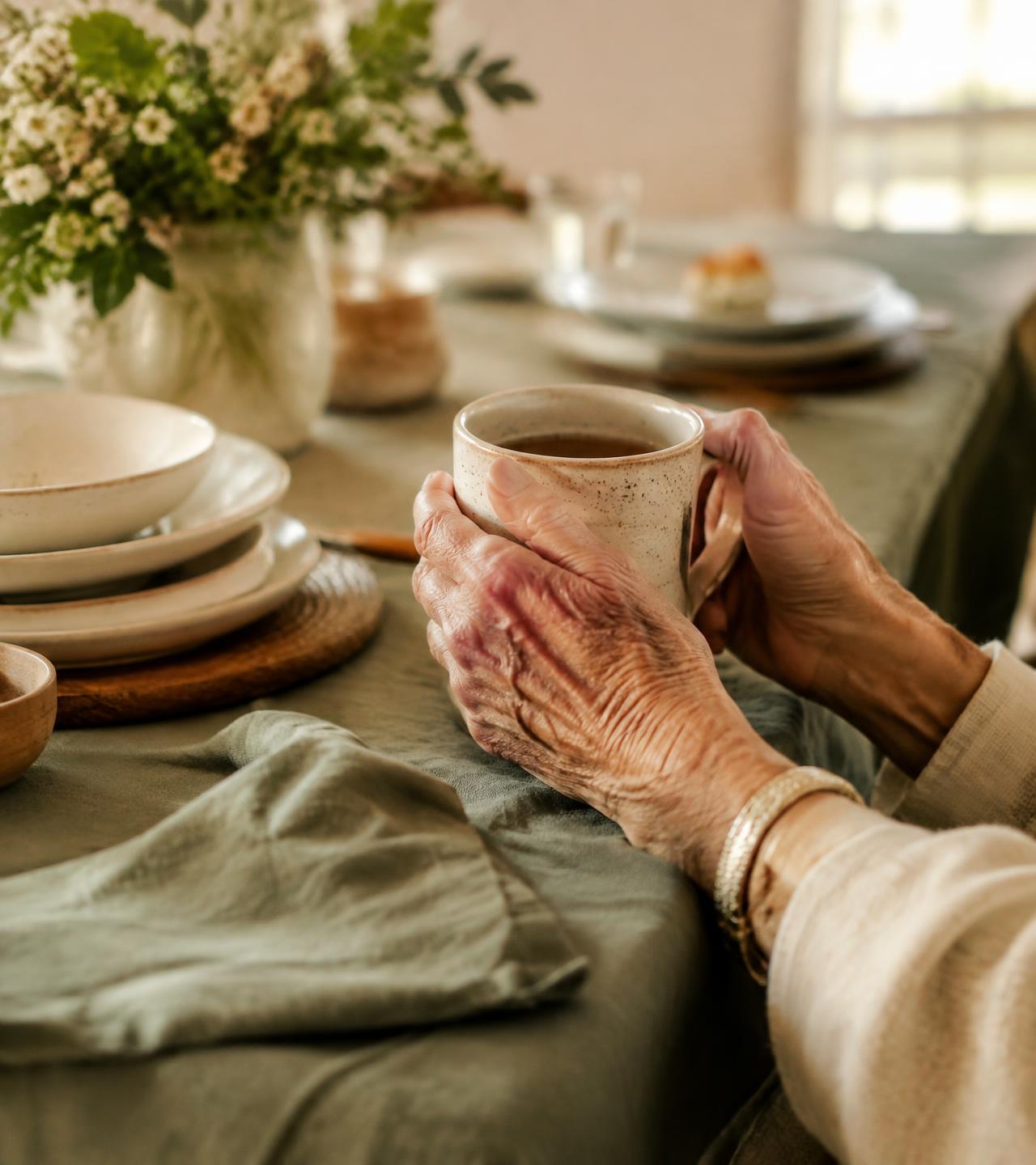Mãos segurando uma caneca de duas alças em uma mesa acolhedora